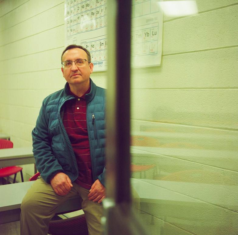 A person sits on a table in a room with white brick walls and periodic table posters, seen through a glass panel with a vertical frame dividing the view.
