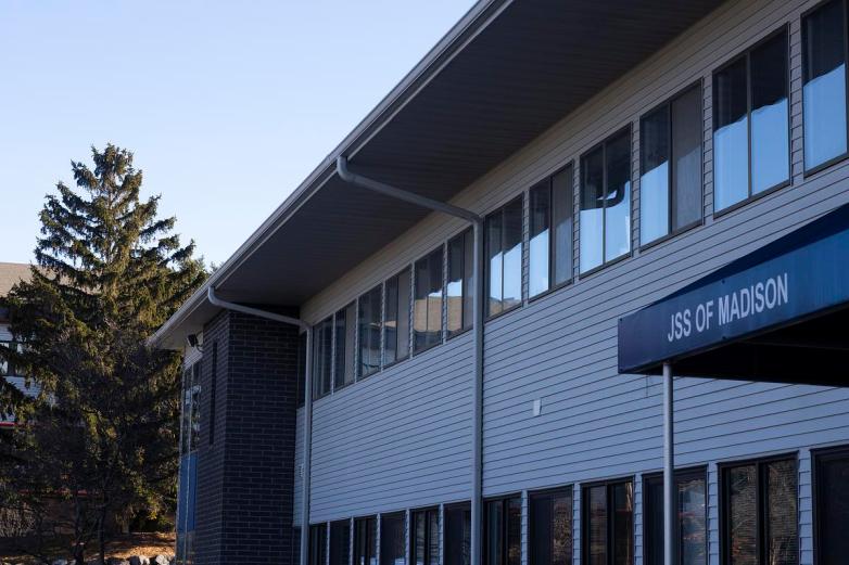 A two-story building with rows of windows displays a sign reading “JSS of Madison” above an entrance, with trees and neighboring buildings nearby.