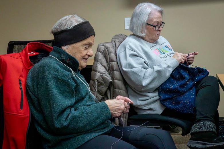 Two people sit in chairs knitting with needles and yarn, with coats draped over the backs of chairs inside a room.