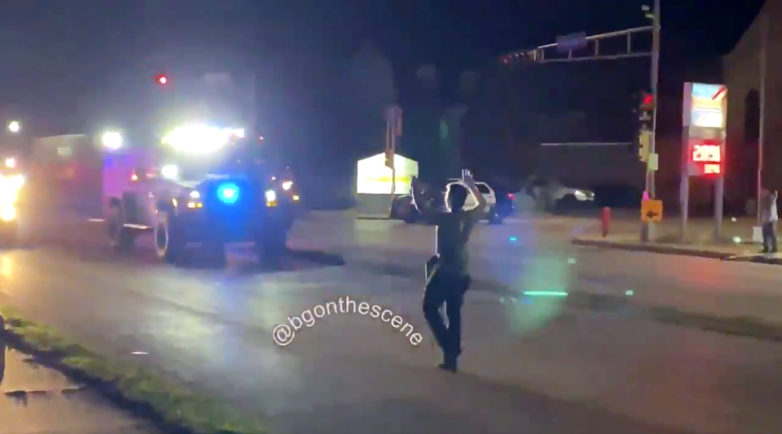 A person stands in a street at night with hands raised, facing a vehicle with flashing lights, while buildings, traffic signals and a few other people are in the background.