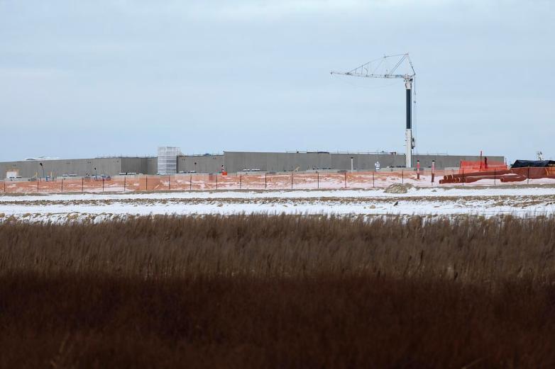 A large industrial building sits behind a fenced construction site with snow-covered ground, orange safety fencing, stacked pipes, and a tall crane rising above the structure.