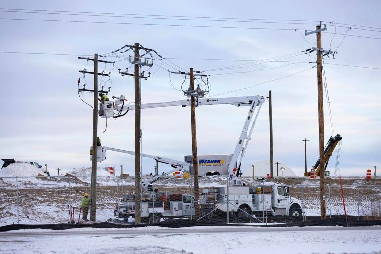 People stand in raised bucket lifts beside wooden utility poles, with power lines overhead and white service trucks parked behind a chain-link fence on snowy ground.