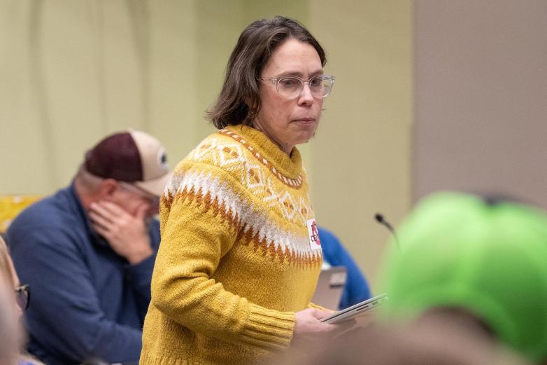 A person wearing a patterned yellow sweater stands holding a tablet, with other seated people and a microphone visible in the background.