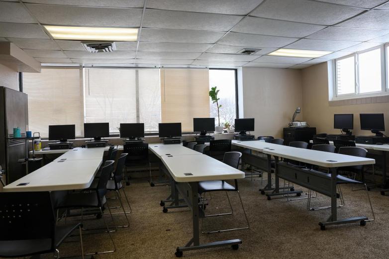 An empty room contains rows of tables and chairs, with computers in rows next to windows with blinds along two walls, and fluorescent ceiling lights.