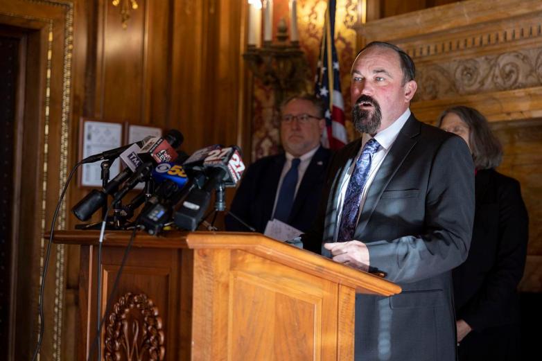 A person stands at a wooden podium speaking into multiple microphones, with other people standing in the background and a U.S. flag visible in an ornate room.