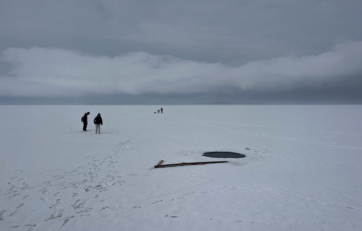 People stand on a snow-covered frozen lake near a round hole in the ice, with wooden planks beside it and footprints across the surface under cloudy skies.