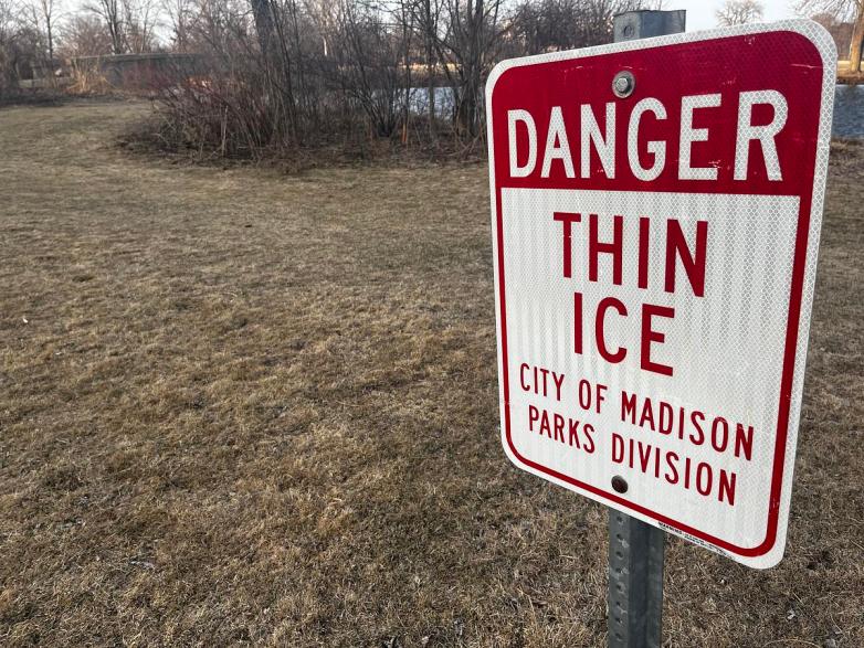 A red and white sign on a metal post reads “DANGER THIN ICE City of Madison Parks Division,” with brown grass, leafless trees, and water in the background.