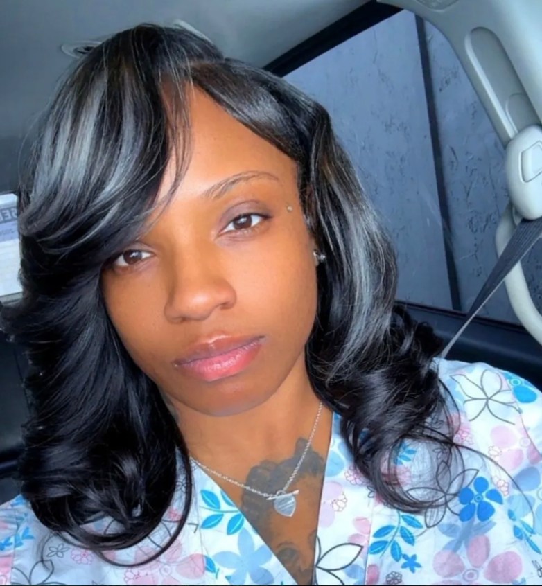 A person faces the camera inside a car, wearing a patterned top and a necklace, with a seat belt visible and daylight coming through the window.