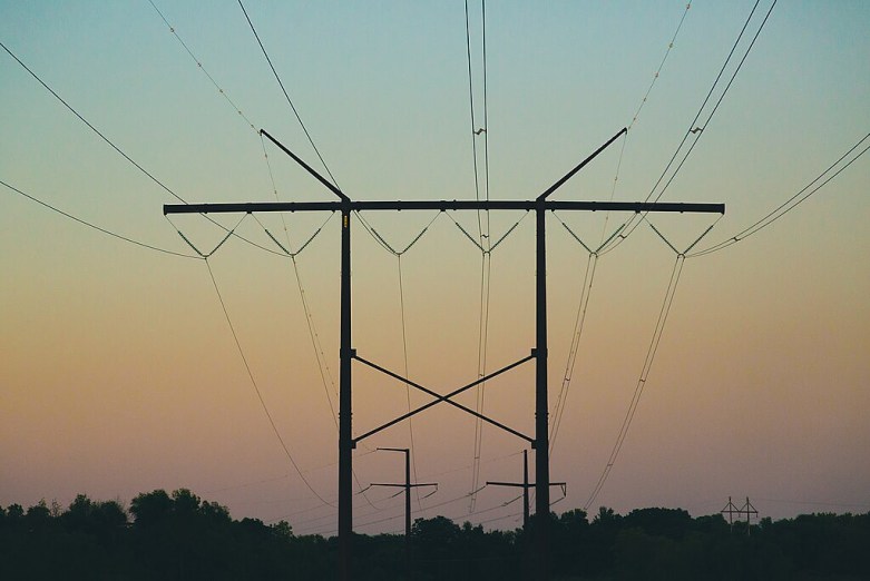 High-voltage transmission towers support multiple power lines stretching across the sky above a tree line at dusk