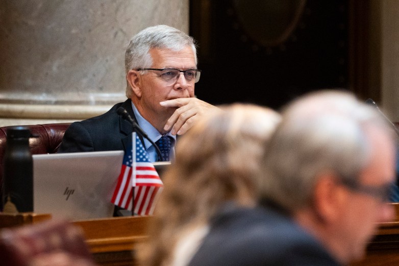 A person sits at a desk with a laptop and a microphone, resting a hand near the mouth, with a small U.S. flag in front and other people blurred in the foreground.