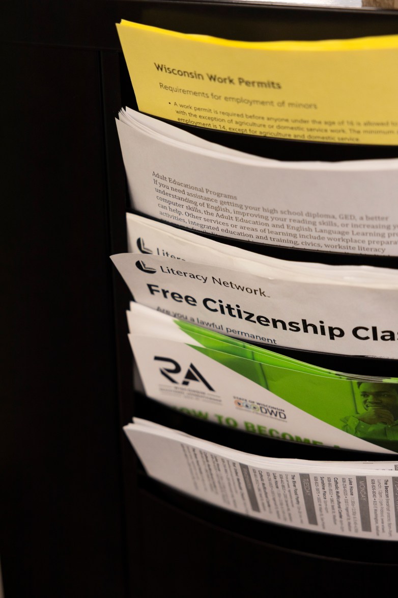 Pamphlets are stacked in a wall rack, with visible titles reading “Wisconsin Work Permits,” “Adult Educational Programs” and “Free Citizenship Class”