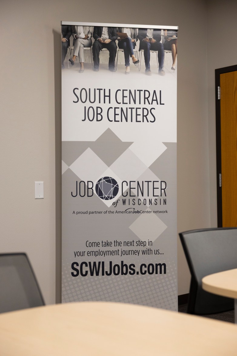A vertical banner indoors reads “SOUTH CENTRAL JOB CENTERS” and “JOB CENTER of WISCONSIN,” with text below saying “Come take the next step in your employment journey with us… SCWIJobs.com”