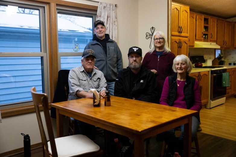 Five people pose around a wooden table near a kitchen, with cabinets, a stove and windows behind them.