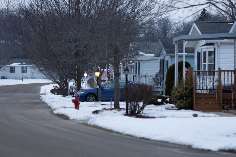 A curving street lined with white homes with snow on lawns and roofs, leafless trees, lit porch lights, a parked blue car, and a red fire hydrant.