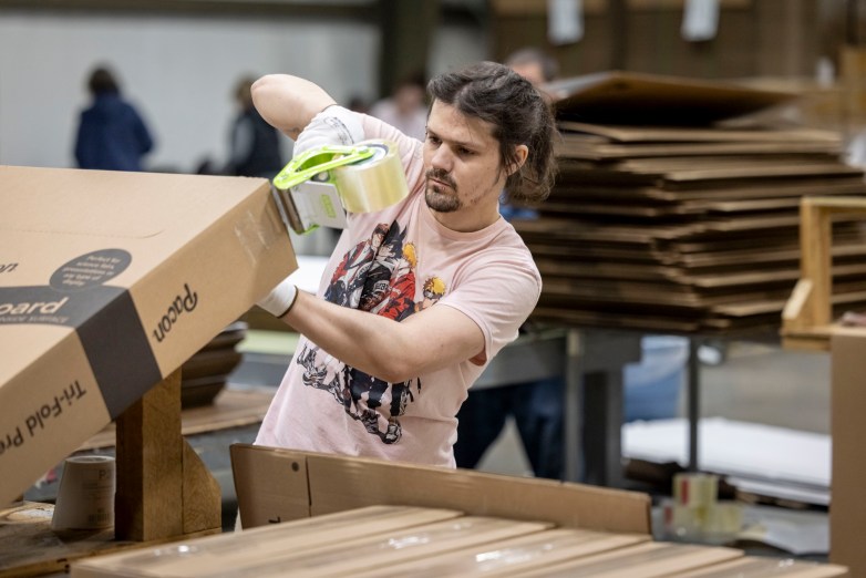 A person uses a tape dispenser to seal a cardboard box at a worktable, with stacks of flattened material piled in the background.