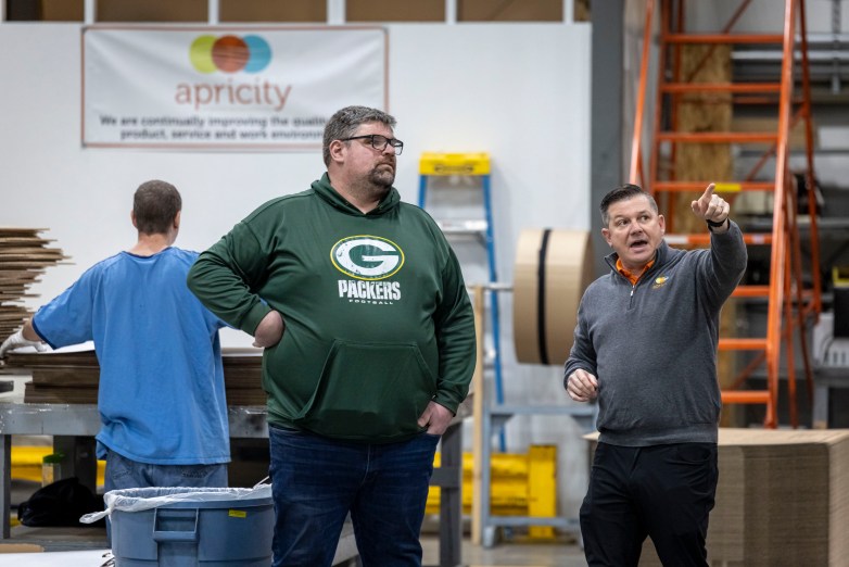 Three people stand in a warehouse-like room — one pointing a finger, one wearing a green Packers sweatshirt and the other facing the other way — near stacked materials and equipment, including a ladder and large rolls, beneath a sign reading