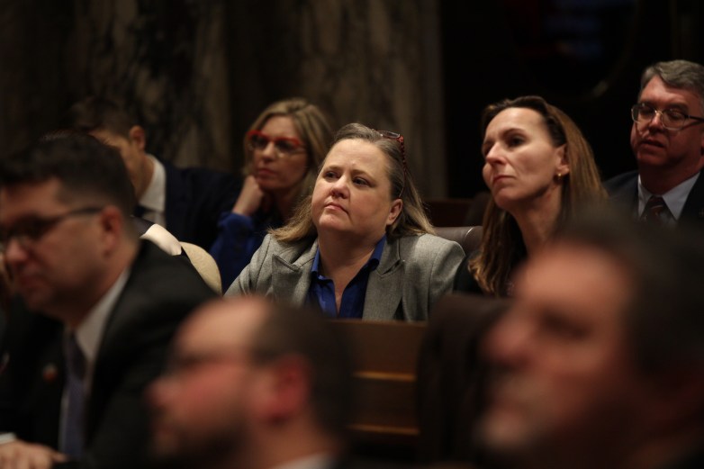 People sit in rows in a room, with a person in a gray jacket and blue shirt centered, while others are seated nearby and blurred in the foreground.