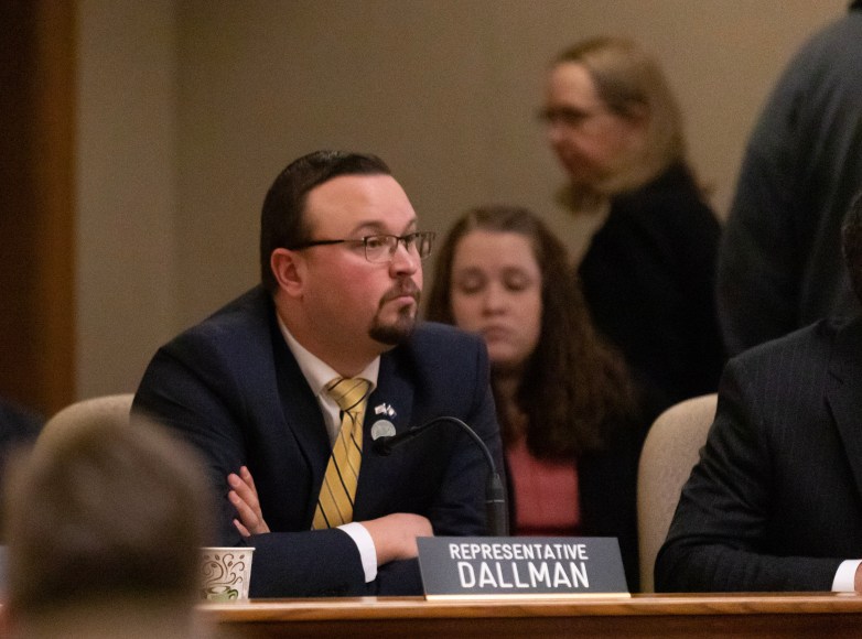 A person sits at a desk with arms crossed, wearing a suit and tie, with a nameplate reading “Representative Dallman” and a microphone in front of the person with other people in the background.