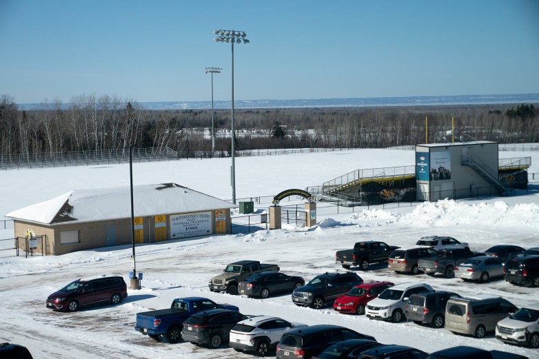 A snow-covered football field and bleachers are behind a parking lot filled with cars. A building next to the football field entrance has a sign that says 