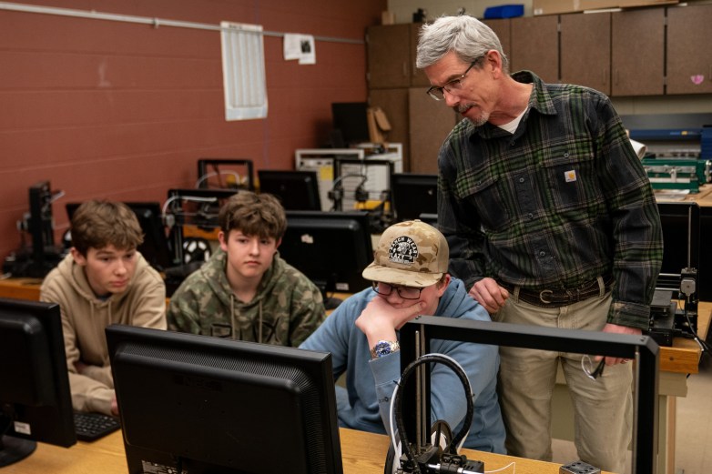 A person stands beside three other people who are seated at a table in a room, looking at a computer monitor, with more computers and other equipment on more tables behind them.