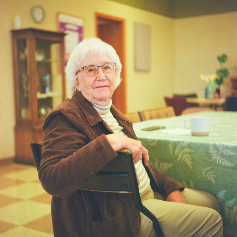 A person with glasses sits at a table with a patterned tablecloth and a mug on it in a room with a cabinet, a clock on a wall, a door and other out-of-focus items in the background.