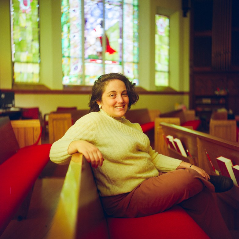 A person sits on a wooden pew inside a church, with red cushions for the pews and stained glass windows in the background.