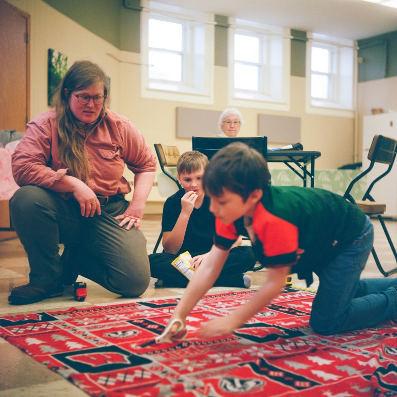 A person kneels beside two children, with one of them holding scissors on a patterned blanket on the floor, while another person sits at a table in the background.