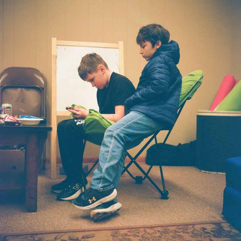 Two people sit on a green folding chair in a room, one looking at a phone while the other leans back, with a small table holding snacks and a whiteboard nearby.