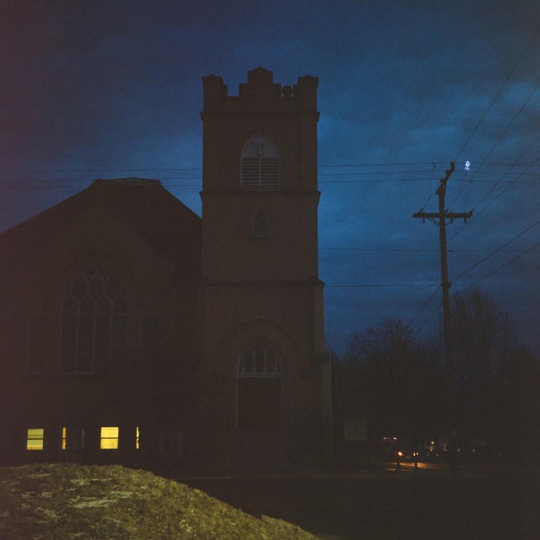 A church building stands in low light with a tower and lit windows, with power lines and a road visible nearby.