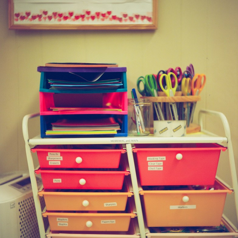 A cart holds drawers with labels for markers, tape, pencils, sharpeners, erasers and other items, with scissors, stacked folders and containers on top of the cart.