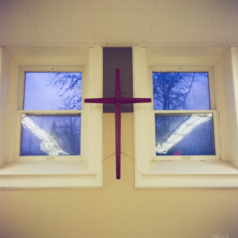 A wooden cross is mounted between two windows showing trees and sky outside.
