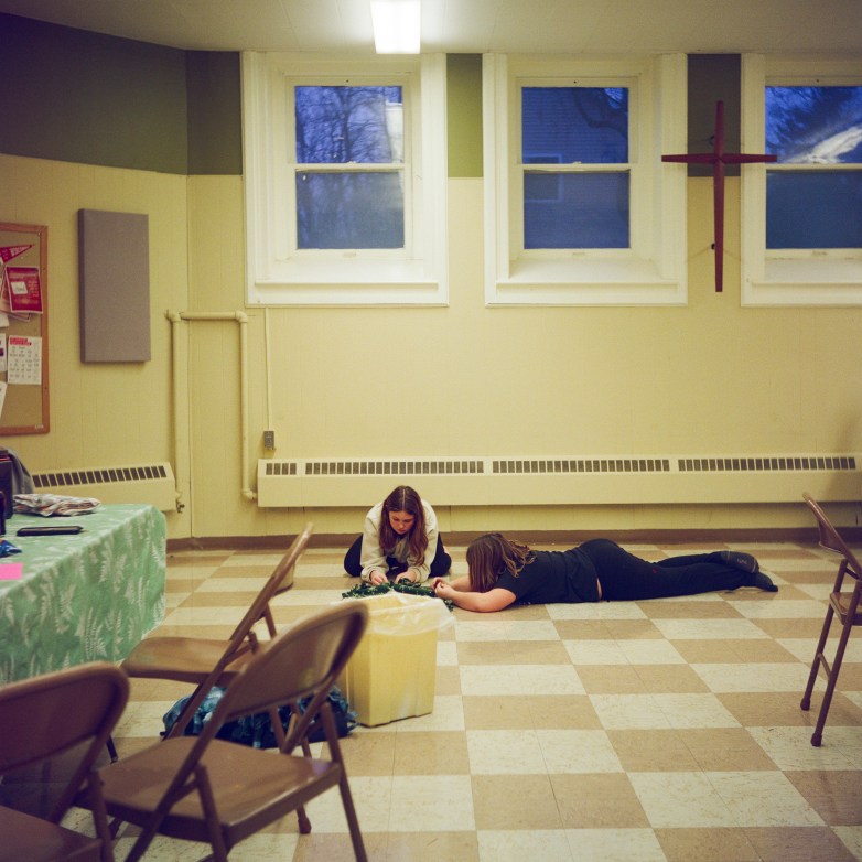 Two people lie and kneel on a tiled floor holding small objects, with folding chairs and a table visible in the room and a wall cross mounted between windows.
