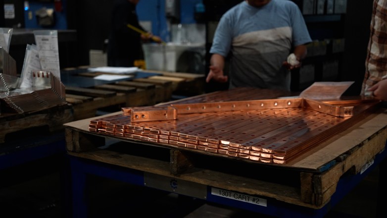 Stacks of copper bars with drilled holes sit on a wooden pallet in a workshop, with a person standing nearby in the background.