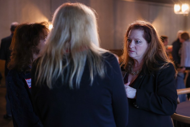 A person faces two others in conversation indoors; one person is seen from behind while another stands nearby in a softly lit room