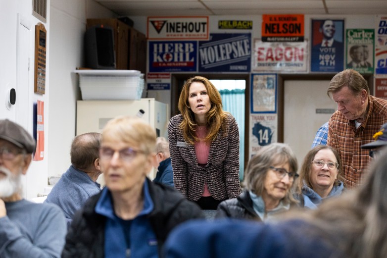 A person leans forward speaking in a crowded room; people sit in chairs around them, and campaign signs line the wall behind including 