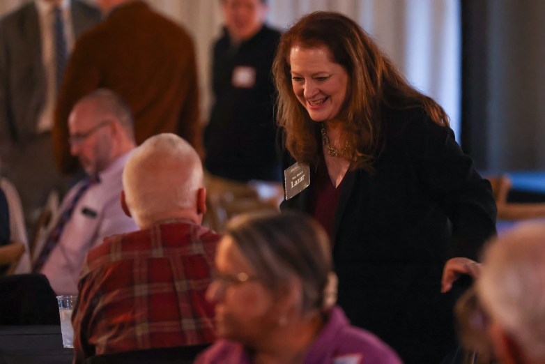 A person leans toward seated people at tables in a softly lit room; others sit and talk while the person engages with someone in a plaid shirt