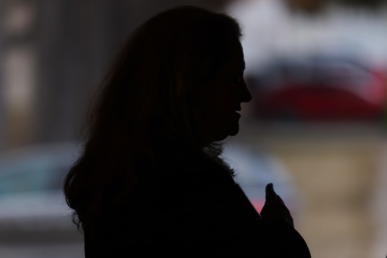 A person appears in silhouette in profile with a hand raised near their chest; blurred shapes of cars and buildings are visible in the background