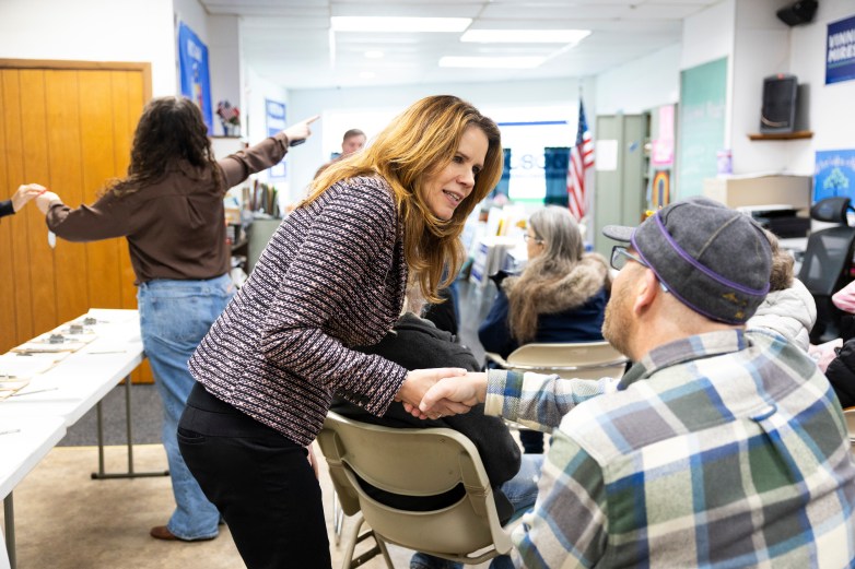 A person leans forward to shake hands with a seated person wearing a cap in a meeting room; others sit on folding chairs and a person gestures near a table in the background