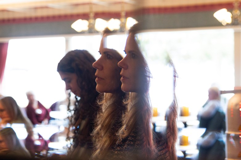 A person’s face appears in profile and reflected in a mirror or glass; other people sit at tables in a bright room with windows in the background