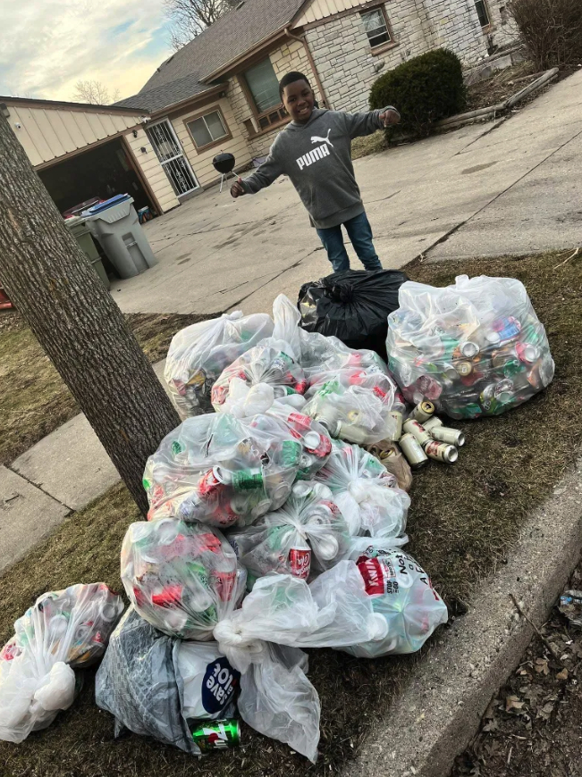 A person stands near a curb beside multiple bags filled with cans and bottles, with a house and driveway in the background.