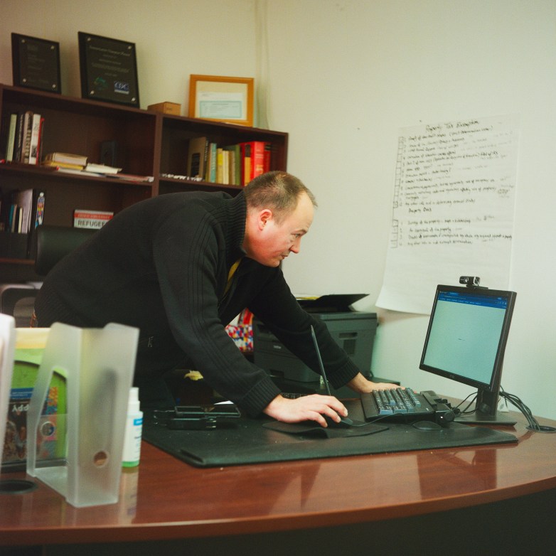 A person leans over a desk holding a computer mouse and looking at a computer screen in a room with books on shelves and a large white piece of paper with words on it on a wall.