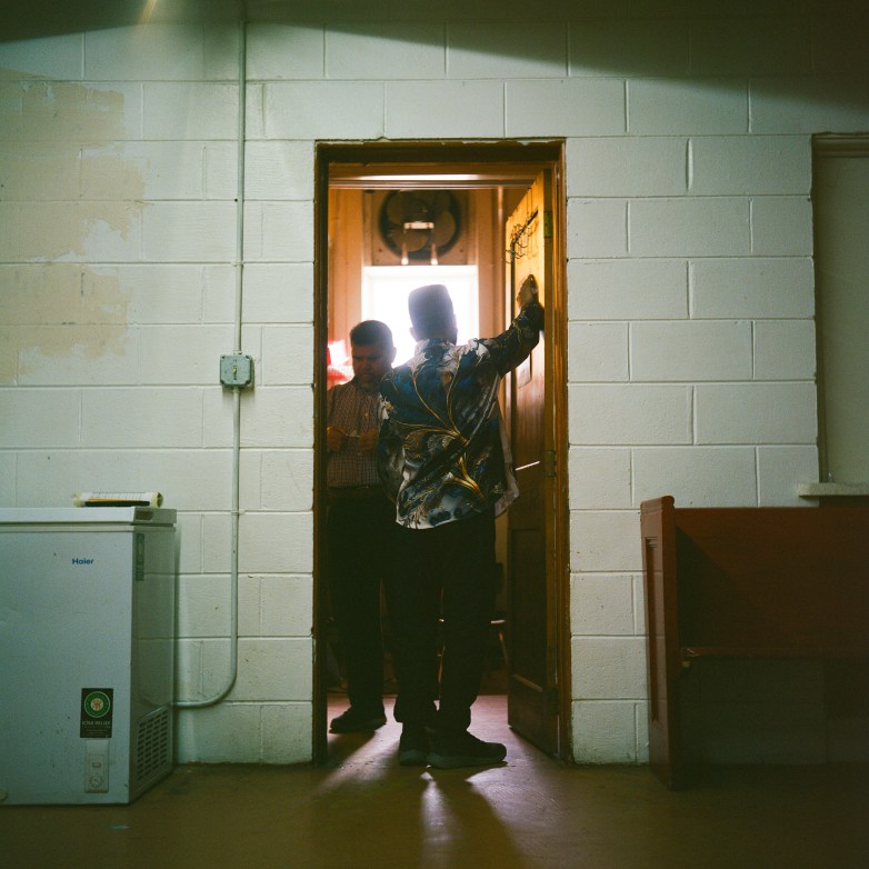 A person stands in a doorway leaning on the door frame while another person stands inside the room, with light shining in from a window behind.