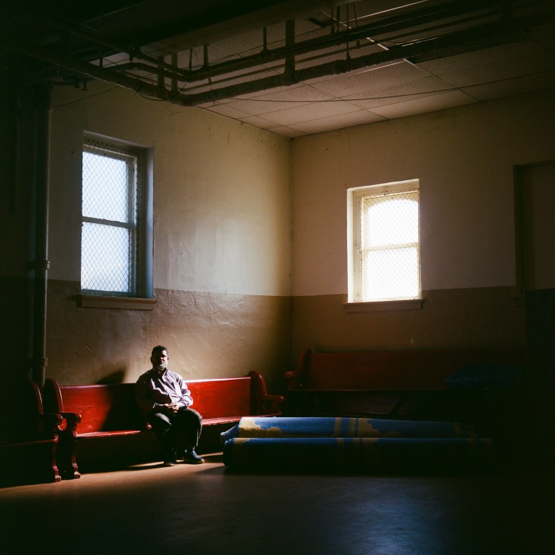 A person sits on a red wooden bench in a room with two windows as light streams in and illuminates part of the floor and nearby rolled rugs.
