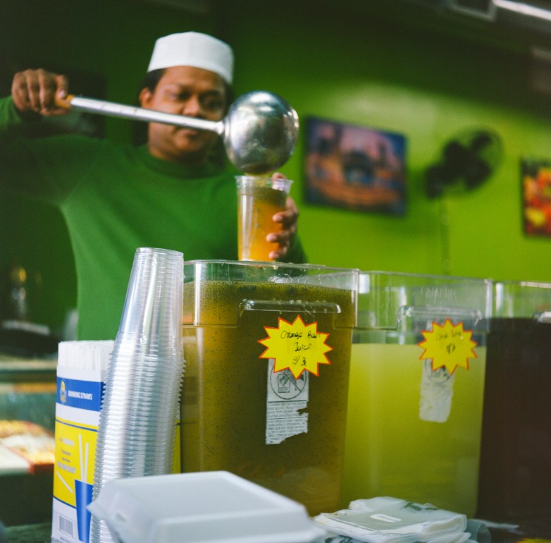 A person pours a drink into a plastic cup from a ladle with a large container labeled 