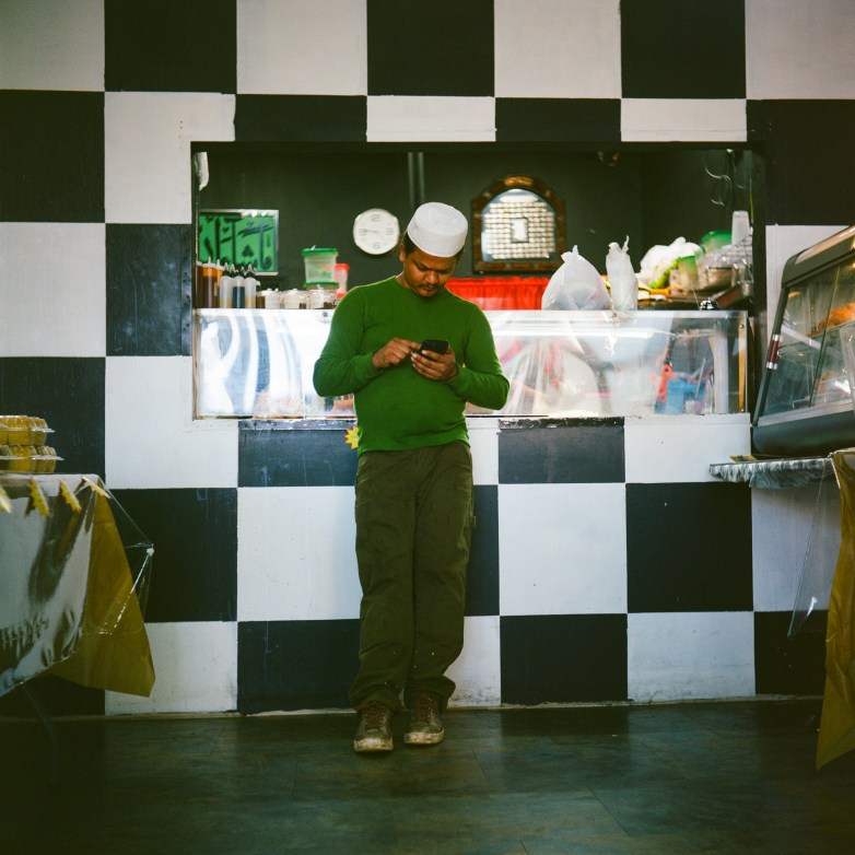 A person in a green shirt and white cap stands and looks at a phone while leaning against a counter with a black-and-white checkered wall.