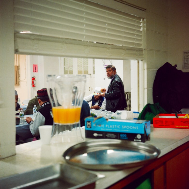 A view through a service window shows people sitting at a table while a person to the right carries a plate, with a pitcher of orange drink and trays on a counter in the foreground.