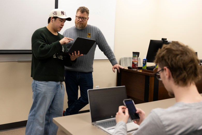 Two people stand in a room looking at a laptop while another person in the foreground sits holding a phone at a desk with a computer on it.