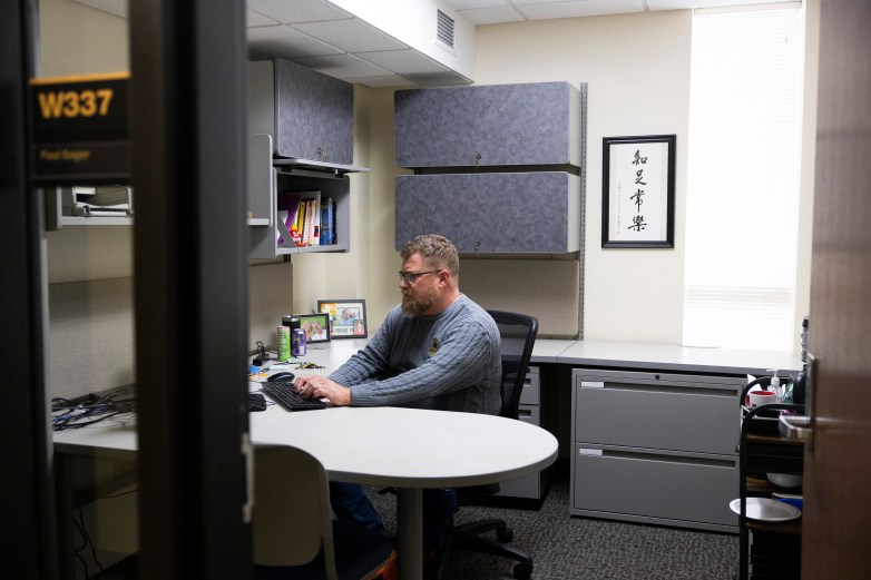 A person sits at a desk with hands on a computer keyboard in a room with shelves, framed photos and a wall hanging.