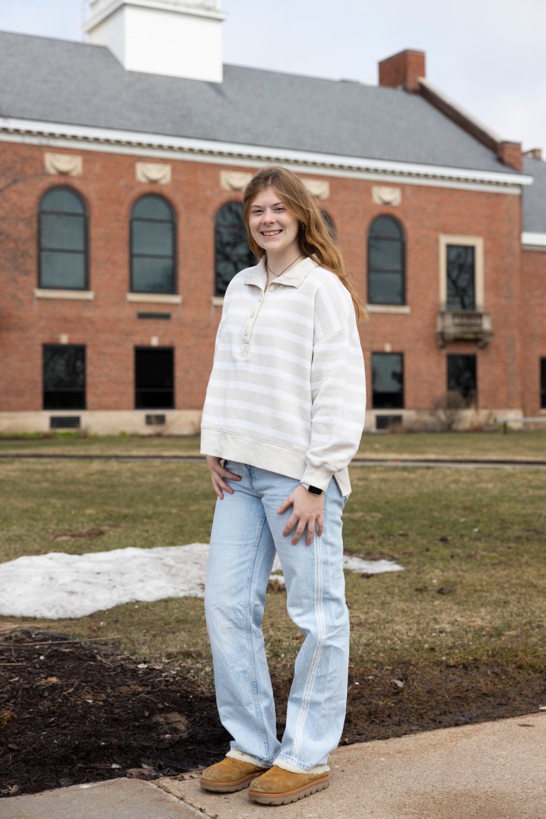 A person stands outdoors in front of a brick building with arched windows, wearing a light sweater and jeans.