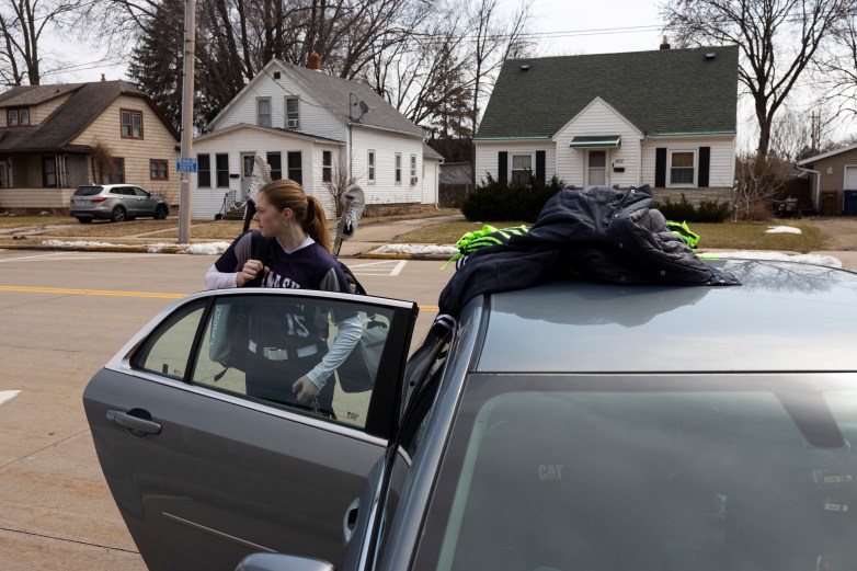 A person in a sports uniform stands beside an open car door holding gear, with jackets piled on the car roof and houses in the background across a street.